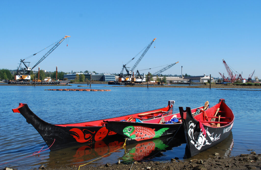 Two Coast Salish canoes on the Duwamish River, Seattle, Washington with an industrial port in the background
