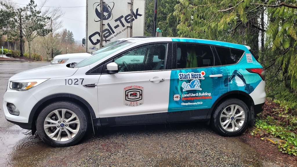 A white car with a blue vinyl wrap image of trees and an excavator behind one logo mark that includes an illustration of a house with trees and test that reads “StartHere!”, and another logo mark with text that reads Land Use& Building Development Basics, KitsapGov.com/DCD