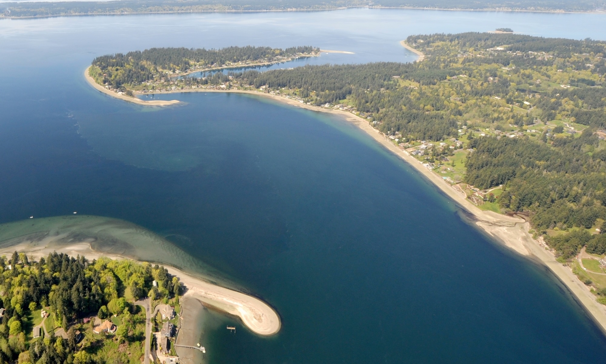Aerial view of Puget Sound showing forested shoreline, sandy beaches, residential areas, and deep blue-green waters with small boats anchored.